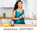 Small photo of Portrait of positive asian woman in nightdress standing at kitchen table with mug of hot drink.