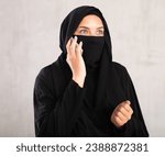 light-skinned Muslim woman with covered face with black niqab is talking on mobile phone. Muslim woman in calm state is talking on smartphone. Close up on gray background