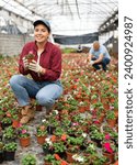Small photo of Young woman gardener holding flower pot with blooming impatiens waller in greenhouse