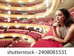 Small photo of Dreamy young adult woman sitting on balcony in theater hall during intermission, examining interior in anticipation of continuation of theatrical production..