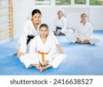 Small photo of Focused sporty young woman in kimono sitting on tatami and stretching in butterfly pose with assistance of trainer pressing on her knees to achieve deeper stretch. Martial arts training routine
