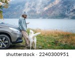 Small photo of Young woman traveler standing next to the car while traveling with her white swiss shepherd dog on the shore of a mountain lake in foggy autumn weather