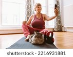 Small photo of Relaxing yoga exercises with pets for overweight adult women with large breasts. Middle-aged, curvaceous, blonde woman with hair tied in bun, sits on gymnastic mat next to dumbbells, takes selfie