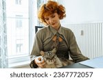 Small photo of Portrait attractive teenage girl with red hair, wearing hipster clothes, shirt and cravat, holding fluffy domestic cat in arms, purring. Home life pets and their owners, real life