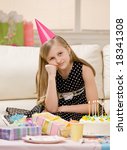 Small photo of Unhappy and ungrateful girl in party hat sits with gifts and birthday cake at party