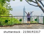 Young female tourist with raised arms to the sky enjoying awesome view of Taipei from top of mountain. Taipei skyline, Taiwan. Wonderful cityscape. Skyscrapers and other modern buildings of downtown.