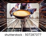 Housewife using dishcloth for taking cheesecake out of oven in kitchen. View from inside of the oven. Woman wearing colorful checkered shirt and black apron.