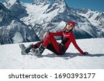 Young pretty fashionable girl in red black color 
ski suit and grey ski boots is sitting on snow and relaxing. She wears fashionable sun glasses, red cap and belt. Background is high snow mountains.