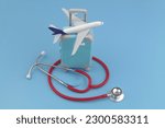 Airplane model on suitcase and red stethoscope on blue background. Travel insurance, medical tourism and healthcare concept.