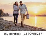 Happy caucasian fashionable couple in love holding hands and walking on coast near river. Man holding picnic basket. In background is sunset.