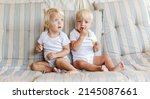 Small photo of Two toddler twins pose for a family photo album. Cute babies in white baby bodysuits eat candy on the sofa and sit next to each other. Spontaneous portrait of twins with blue eyes and blonde hair