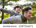 young asian father carrying son on back having fun enjoying nature outdoors in park