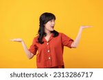 Portrait beautiful young asian woman happy smile dressed in orange clothes showing hand gesture, arm compare two variants demonstrate empty space isolated on yellow studio background.
