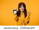 Small photo of Punctuality meets style as young Asian woman, donning a yellow sweater and red beret, flashes a happy smile beside her trusty alarm clock in her stylish workspace.