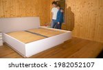 Small photo of CLOSE UP: Young Caucasian couple hugs after assembling their king sized bed in the wooden bedroom. Man and woman mount bunkie boards onto the bed frame of a large bed in their brand new bedroom.