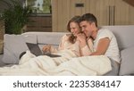 Small photo of CLOSE UP: Couple holding hands while watching their photos and videos on laptop. Loving couple sitting on couch under blanket looking at laptop screen. Twosome enjoying at home on a cold winter day.