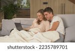 Small photo of CLOSE UP: Young married couple snuggling on couch and watching photos on laptop. Loving couple sitting on couch under blanket looking at laptop screen. Twosome enjoying at home on a cold winter day.