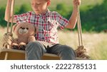 Small photo of Happy boy is swinging on swing in park with his teddy bear toy. Son is swinging on swing in park, kid smiling. Child plays on wooden swing, childish loneliness, soft toy bear friend. Dream concept.