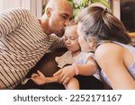 Small photo of Dad and mom smooch their baby girl while she eats baby food with spoon, loving family on sofa in living room