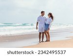 Small photo of Happy couple in love on the beach vacation walking together at sunset. Young woman in white dress and caucasian man in white shorts.