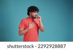 Small photo of Young latin spanish man in coral t-shirt on blue studio background, scarf sneezing or coughing covering mouth with hands.