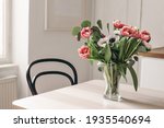 Spring flowers in glass vase on wooden table. Blurred kitchen background with old chair. Bouquet of red tulips, white anemone flowers and eucalyptus branches. Contemporary elegant scandi interior.