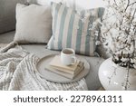 Small photo of Spring interior still life. Cup of coffee, tea on pile of books. Round beige table. Blossoming cherry plum tree branches in ceramic vase. Cozy linen sofa, cushions. Blurred background. Home decor. Top