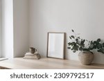 Small photo of Cup of coffee, books. Empty vertical picture frame mockup on wooden desk, table. Vase with silver eucalyptus tree branches. Minimal working space, home office. Scandinavian interior design. Side view.