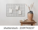 Small photo of Black metal mesh noticeboard, bulletin board with blank memo cards mockups. Elegant home office interior concept. Clay vase with dry grass and cup of coffe on wooden table. White wall background.