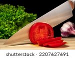A metal knife slicing a red ripe tomato on a wooden board. Close up of a sharp knife blade cutting tomato on slices, green salad and garlic on a black isolated background.