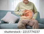 Small photo of Senior Asian man sitting on sofa having suffering from stomach ache holding his stomach pain, Unhappy old aged man feeling discomfort because food poisoning, people health stomachache problem