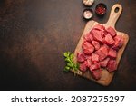 Raw beef meat chopped in cubes with bunch of fresh parsley on wooden cutting board for cooking stew or other meat dish on brown dark stone concrete background top view flat lay space for text