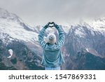 Back view of young traveler girl in gloves and hat standing over snowy mountain peaks and making shape of love heart by hands. Winter travelling scene, wanderlust concept.