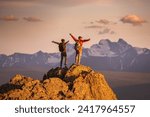 Two happy young tourists with backpacks are standing with open arms at mountain top and enjoys sunset view