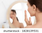 Closeup of unrecognizable lady using face cream at home, looking at mirror and smiling. Young woman applying beauty product on her cheeks, nourishing her sensitive skin after shower, face care
