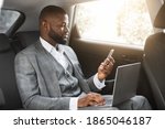 Handsome african american businessman with laptop holding smartphone while going by car on business trip, copy space. Black entrepreneur looking at mobile phone screen, using laptop