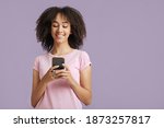 Modern devices and online communication. Happy young african american lady in pink t-shirt with curly hair looking in smartphone and blogging isolated on purple background, empty space, studio shot