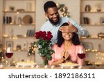 Small photo of Greeting With Holidays. Smiling black man covering his woman eyes and giving her bunch of red roses, making surprise to beautiful lady. African american couple celebrating together at home or cafe