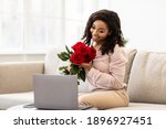 Holidays And Gift Delivery. Happy Black Young Woman Holding Bouquet Of Red Roses And Showing To Webcam, Sitting On Couch At Home And Using Laptop, Having Video Call. Virtual Celebration Concept