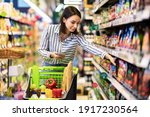 Portrait Of Millennial Lady Holding And Using Smartphone Buying Food Groceries Walking In Supermarket With Trolley Cart. Female Customer Shopping With Checklist, Taking Products From Shelf At The Shop