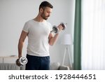 Closeup of handsome middle-aged man doing dumbbell workout at home, working on arms strength, looking at his biceps, copy space. Athletic man lifting dumbells up over living room interior