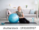 Full length portrait of happy mature woman resting on yoga mat with fitness ball, smiling at camera indoors, free space. Cheerful senior lady taking break from her domestic training