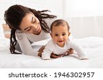 African Mother Playing With Cute Baby Daughter Helping Her Crawl On Bed In Bedroom At Home. Young Mommy Enjoying Motherhood Routine And Child Care. Maternity Leave Lifestyle