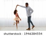 Small photo of Handsome black father and little daughter in beautiful dress dancing waltz at home, standing next to window, full length photo. Loving african american dad and school girl having fun together at home