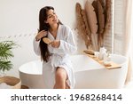 Haircare. Brunette Female Brushing Hair With Hairbrush Detangling Split Ends Smiling Sitting On Bathtub Wearing Bathrobe In Modern Bathroom At Home. Beauty Routine Concept