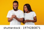 Small photo of Portrait of shocked African American woman spying on her smiling boyfriend who using mobile phone, texting sms or scrolling social media news feed. Black couple standing over yellow studio background
