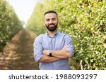 Confident owner of eco farm, work outside, successful business and growing orchard. Friendly smiling bearded young man with crossed arms on chest on path on plantation with green apple trees in garden