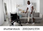 Small photo of Surgery rehabilitation of bone and orthopaedic recovery, stay at home. Smiling adult african american guy with broken leg in cast walks with crutches to wheelchair in living room interior, panorama