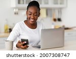 Business And Telecommuting Concept. Portrait of smiling young black woman using her smartphone and modern laptop computer, reading text message, sitting at desk with gadgets, working at home office
