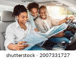 Cheerful African American Family Looking At Road Map Sitting In Car. Parents And Daughter Choosing Destination For Summer Road Trip Together. Local Tourism Concept. Selective Focus
